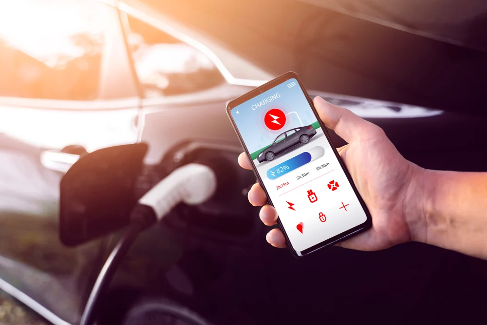 A closeup of a hand holding a smartphone and showing some enabled smart charging features. In the background, a car is plugged in and charging.