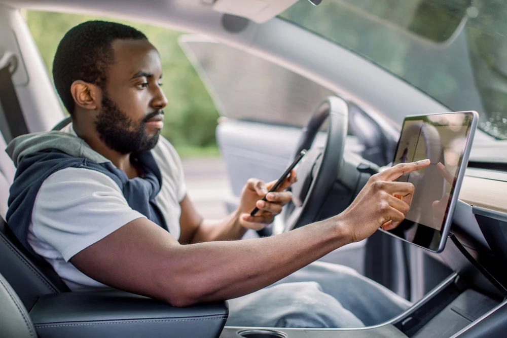A man seated in an electric vehicle is adjusting settings on the dashboard while holding his smartphone.