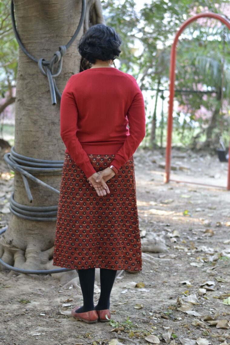 Back view of a woman standing wearing a red Garibaldi Skirt, a red button-up cardigan, and red flats