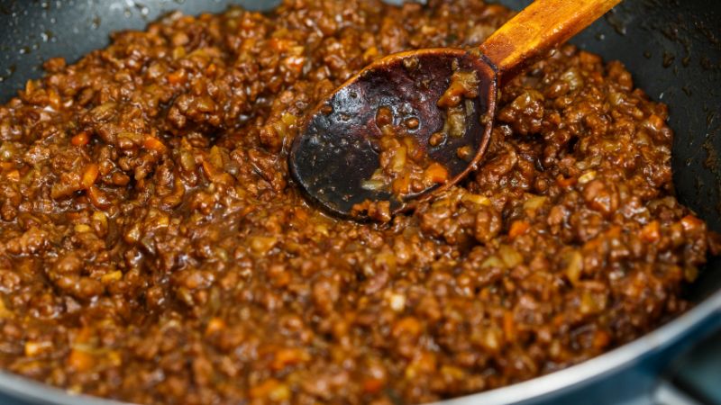 A close-up of cooked minced meat sauce mixed with veggies, stirred with a wooden spoon.