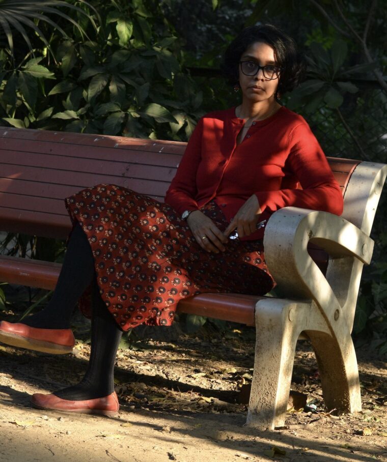 A woman seated on a bench wearing a red Garibaldi Skirt, a red button-up cardigan, and red flats