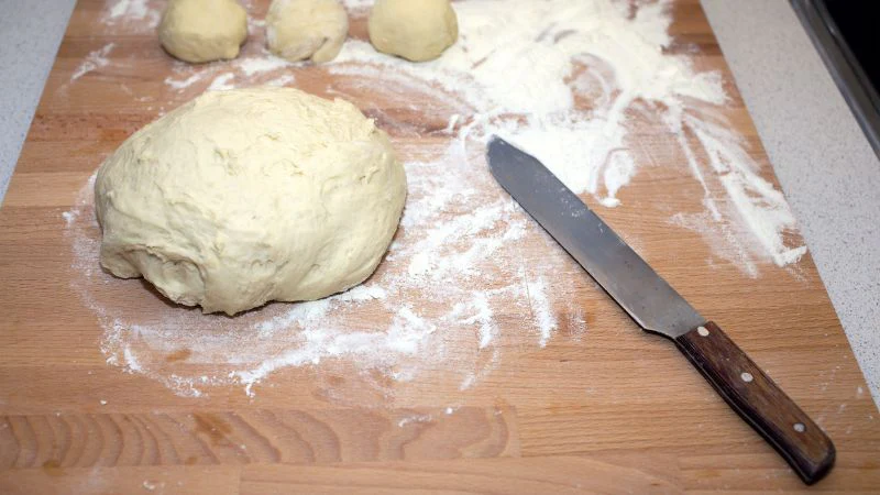 A board with a large dough ball, three smaller pieces, scattered flour, and a knife.