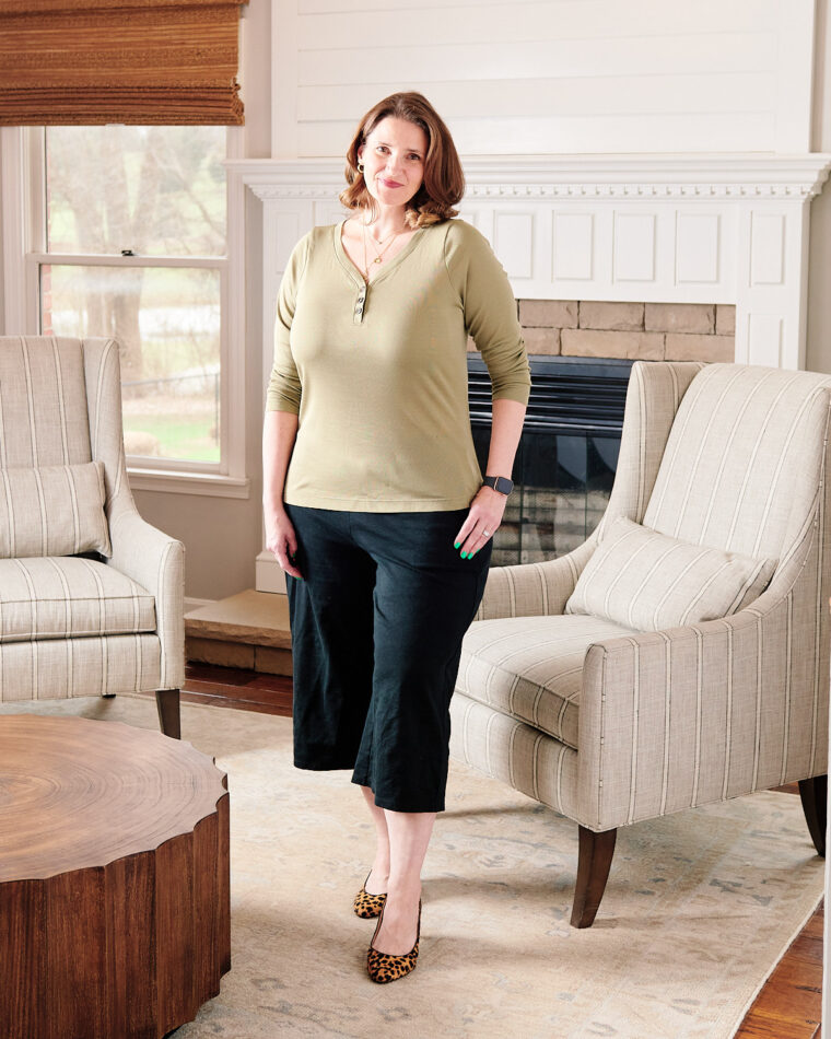 A woman smiling and standing near a fireplace wearing an olive Henley, black cropped pants, and leopard-print pumps.