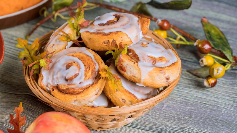 A wicker basket with six cinnamon rolls, white icing, autumn decor, and a peach.
