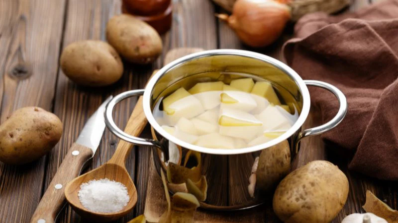 A pot with peeled potato chunks in water, surrounded by whole potatoes, knife, spoon with salt on a brown cloth-covered table.
