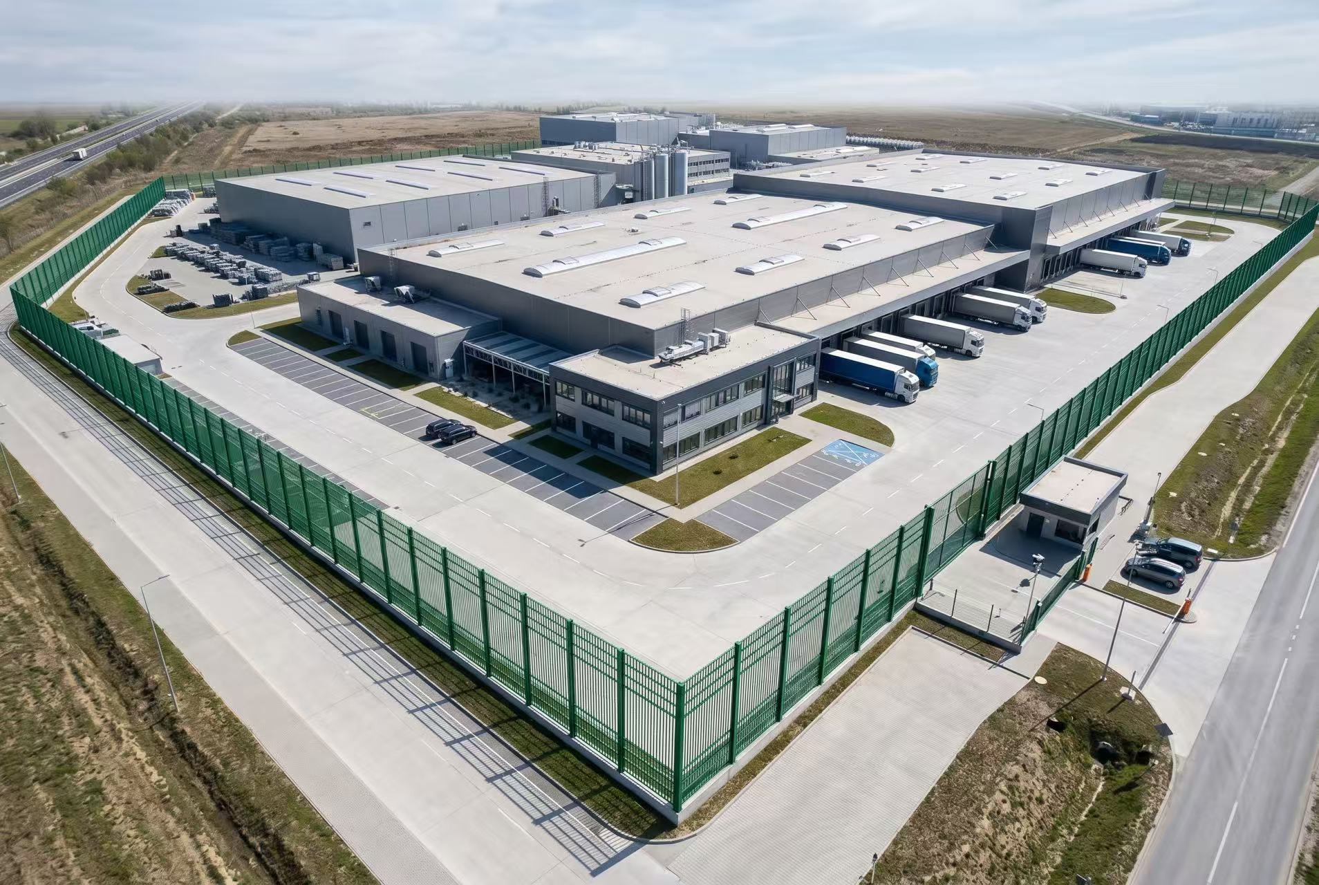 Aerial view of a large industrial facility perimeter enclosed by green powder coated 3D welded wire mesh fence, showing clean uniform color and professional installation quality